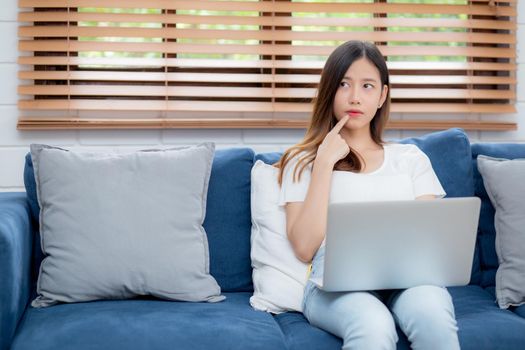 Beautiful young asian woman working on laptop computer and thinking idea on sofa at home, freelance girl sitting on couch using notebook to internet at living room, one person, lifestyle concept.