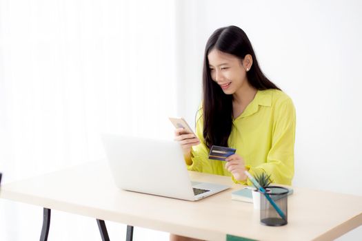 Young asian business woman using smart phone and holding credit card while online shopping and payment online with laptop computer on desk at home, female holding debit card, communication concept.