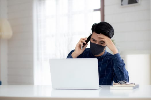 Young asian business man in face mask talking smartphone and work on laptop computer for protect covid-19, businessman quarantine work from home with notebook, social distancing and stay home.