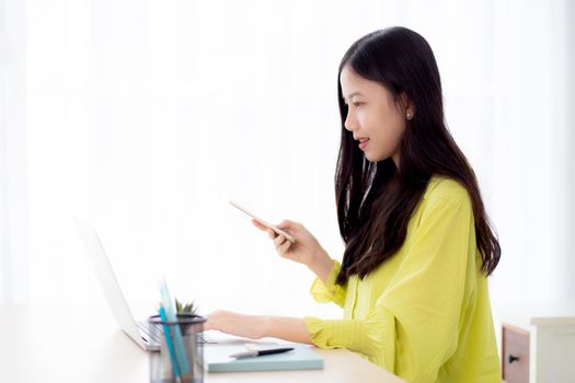 Young asian businesswoman working on laptop computer online on desk at home office, freelance woman looking message on smart phone and typing on notebook on table, business and communication concept.