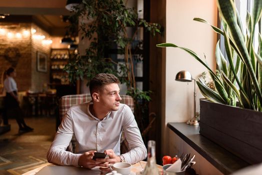 Waist up of guy sitting at the table while holding smartphone in coffee shop. Modern lifestyle concept