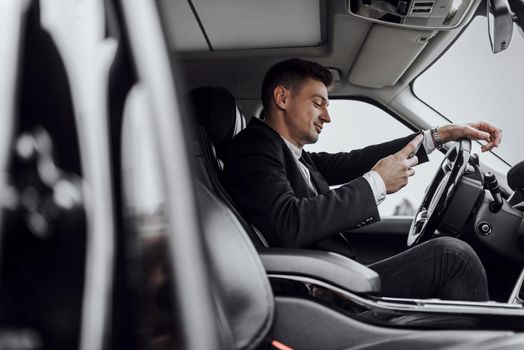 Side view of smiling handsome young man reading message on his smartphone while driving automobile. Rent and trade-in concept