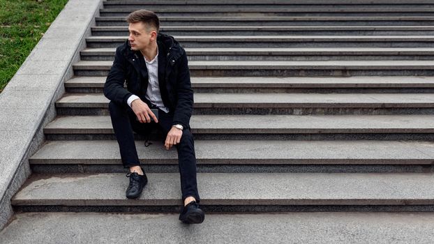 Guy in black jacket sitting on stairs while looking away outdoors. Copy space. Lifestyle concept