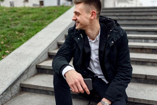 Cropped photo of smiling handsome guy resting on stairs while looking away and holding smartphone. Lifestyle concept