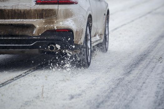 Road snow pieces flow from wheels of dirty white car moving fast in daylight city with selective focus. Car moving on snowy road after heavy winter snowfall.