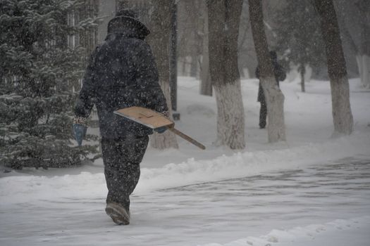 A working man of the municipal service with a snow shovel walks along the road in a storm, blizzard or snowfall in winter in bad weather in the city.Extreme winter weather conditions in the north.