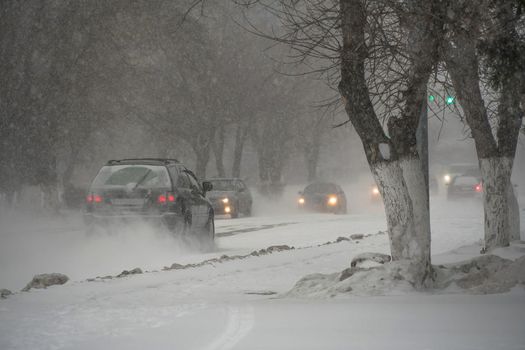 Snow-covered road with cars in a storm,blizzard or snowfall in winter in bad weather in the city.Extreme winter weather conditions in the north.Cars drive through the snow-covered streets of the city