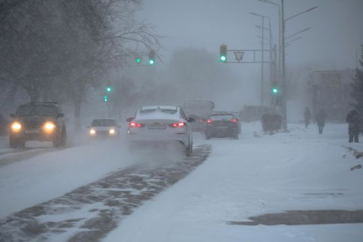 Snow-covered road with cars in a storm,blizzard or snowfall in winter in bad weather in the city.Extreme winter weather conditions in the north.Cars drive through the snow-covered streets of the city