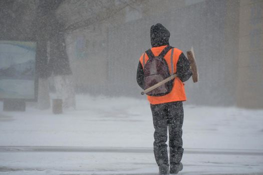 A working man of the municipal service with a snow shovel walks along the road in a storm, blizzard or snowfall in winter in bad weather in the city.Extreme winter weather conditions in the north.