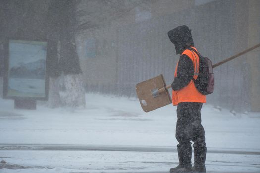 A working man of the municipal service with a snow shovel walks along the road in a storm, blizzard or snowfall in winter in bad weather in the city.Extreme winter weather conditions in the north.