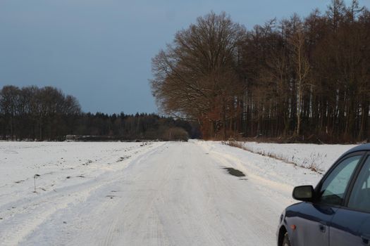 Blue car on snowy dirt road next to a field with trees in the background