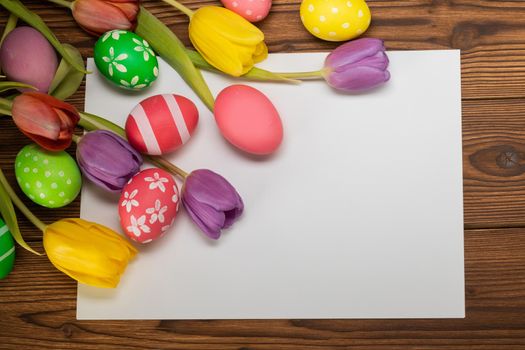 Hand-painted easter eggs with tulips on wooden background and paper copy space for text top view template