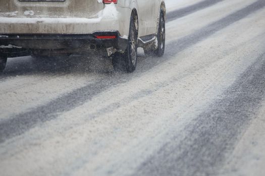 Road snow pieces flow from wheels of dirty white car moving fast in daylight city with selective focus. Car moving on snowy road after heavy winter snowfall.