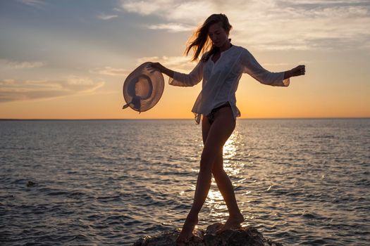 Silhouette of young happy carefree woman posing with hat on the beach at sunset