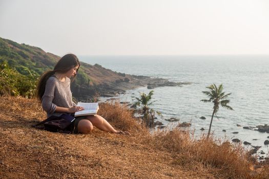 Attractive Asian woman is sitting on the edge of the mountain with a sea view, reading a book. Overall plan