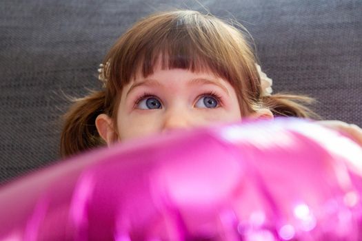 A cute, brown-haired, blue-eyed baby girl sitting on a sofa and hiding her face behind a big pink balloon.