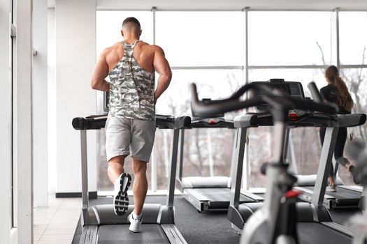 Muscular athletic bodybuilder fitness model running treadmill gym near big window jogging stationary machine do cardio training Weight loss Healthy lifestyle concept. Back view rear full length