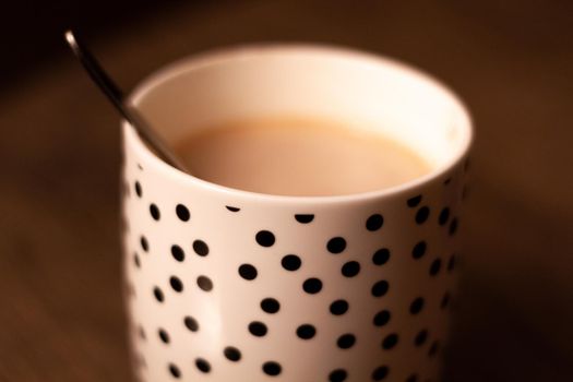 Close-up of a white cup with black dots with a hot drink and a metal spoon on a wood table