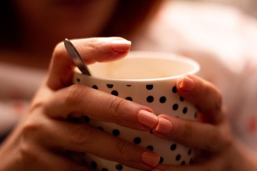 Detail of the hands of a woman holding a white porcelain cup with black dots containing a hot drink