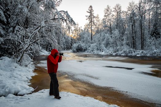 The photographer does to a photo the wild nature at sunset, the wild frozen small river in the winter wood, the Red River, ice, snow-covered trees. High quality photo