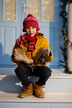 A handsome boy in bright clothes sits on the porch of the house and holds old skates.