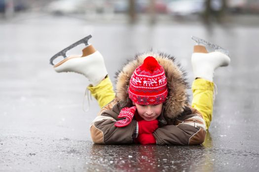 A child in figure skates in winter lies on the ice and smiles.