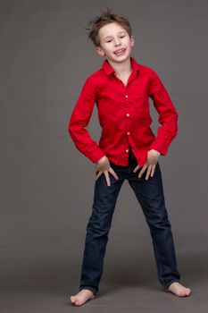 Handsome boy in a red shirt and jeans on a gray background. A child in the modeling business is posing.