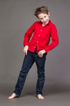 Handsome boy in a red shirt and jeans on a gray background. A child in the modeling business is posing.