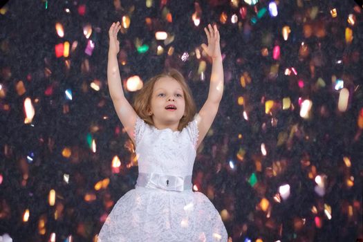 Beautiful little girl in a white dress with raised hands under bright confetti.