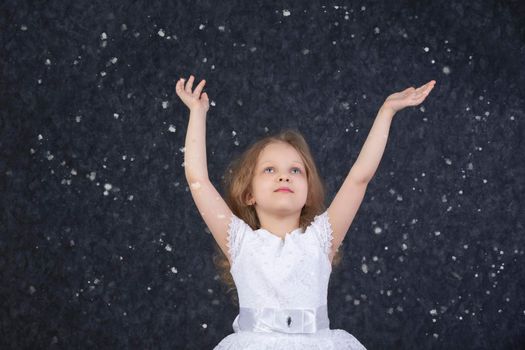 Beautiful little girl in a white dress with raised arms under flying snowflakes.