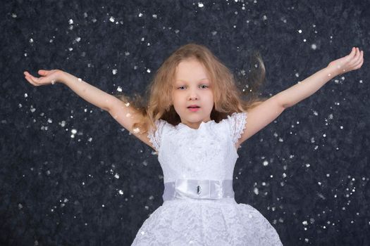 Beautiful little girl in a white dress with raised arms under flying snowflakes.