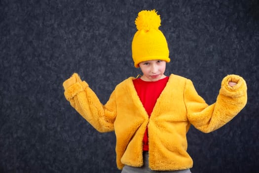 Funny little boy in a fluffy yellow fur coat and a knitted hat on a gray background.