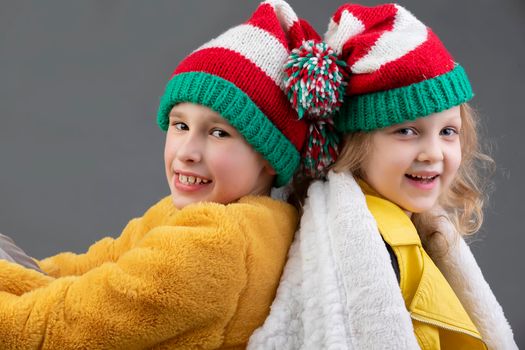 Beautiful little girl and boy in knitted Christmas hats are sitting with their backs to each other and smiling on a gray background. Happy Christmas kids.