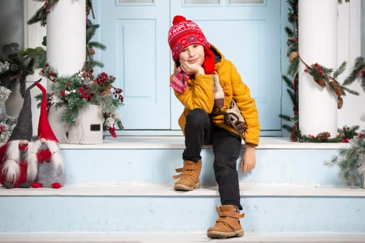Handsome little boy in christmas. Funny child holds old skates and sits on the porch of a house decorated with Christmas tree decorations