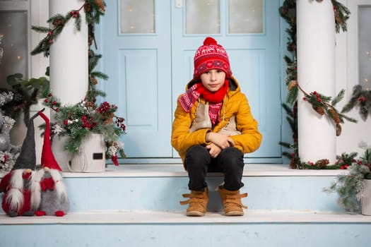 Handsome little boy in christmas. Funny child holds old skates and sits on the porch of a house decorated with Christmas tree decorations