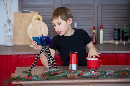 A portrait of a confident and creative child blogger talking to the camera at home in the kitchen, he is vlogging. A little boy is recording a stream on a smartphone.