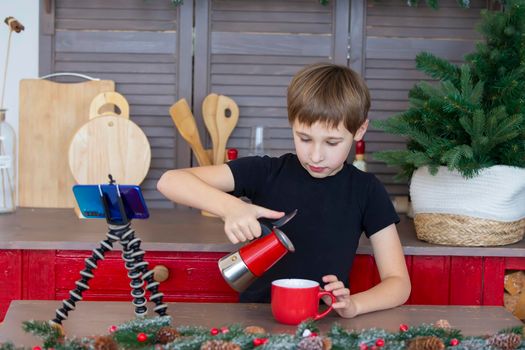 A portrait of a confident and creative child blogger talking to the camera at home in the kitchen, he is vlogging. A little boy is recording a stream on a smartphone.