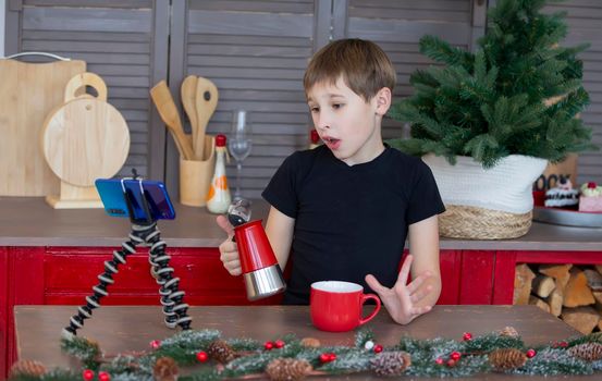 A portrait of a confident and creative child blogger talking to the camera at home in the kitchen, he is vlogging. A little boy is recording a stream on a smartphone.