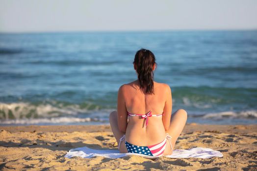 A middle-aged woman sits on the coast in a swimsuit with an American flag print.