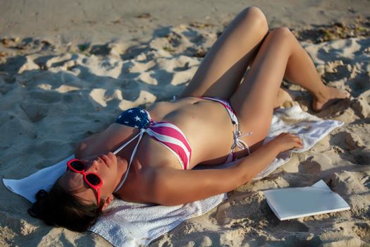 A middle-aged woman lies and sunbathes on the sand in a swimsuit with an American flag print.