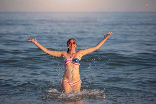 Happy middle aged woman splashing sea water in swimsuit with American flag print.