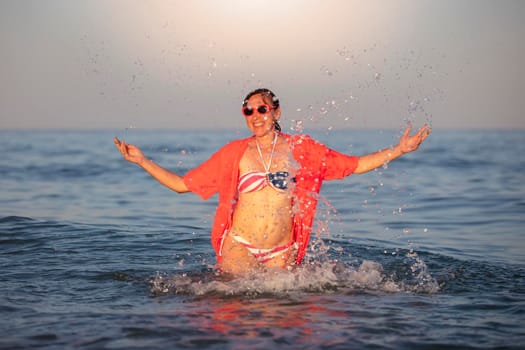 Happy middle-aged woman stands in sea water with raised arms in a swimsuit with an American flag print.