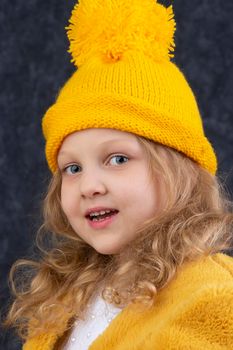 A beautiful little blonde in a knitted yellow hat looks at the camera and smiles. Happy beautiful child about five years old.