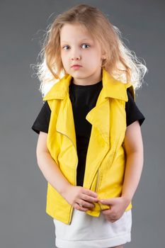 Beautiful little blonde girl in a yellow jacket on a gray background. Five year old girl. Model tests for a child.