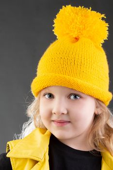 Beautiful little blonde girl in a yellow knitted hat on a gray background.