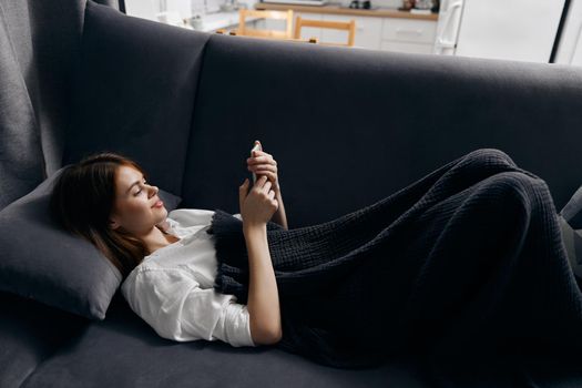 woman resting on the couch with a phone in her hand and a gray plaid on her legs. High quality photo