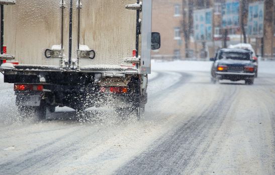 Road snow pieces flow from wheels of dirty truck moving fast in daylight city with selective focus. The truck is moving on snowy road after heavy winter snowfall.