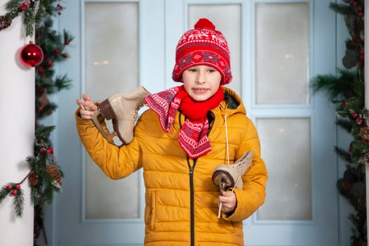 Handsome little boy in bright clothes holds old skates against the background of Christmas decorations.