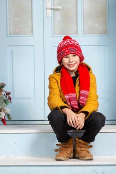 Handsome little boy in bright winter clothes sits on the porch of the house. Happy child in winter.