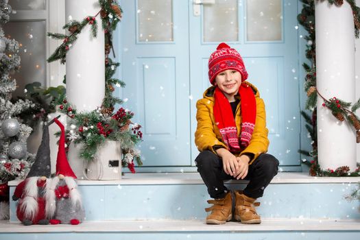 A handsome little boy in bright clothes sits on the porch of a house decorated with Christmas decorations.